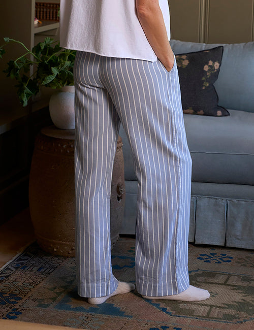 A woman stands in a living room wearing a white short-sleeve t-shirt and light blue and white vertically striped pajama pants, viewed from the back., alternate view