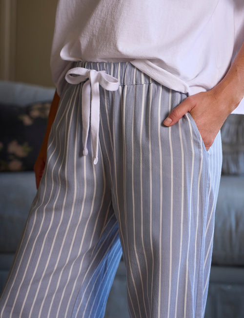 Close-up of a person wearing loose, wide-leg pants with vertical light blue and white stripes, a white drawstring waist, and side pockets, paired with a white top in an indoor setting., view 3