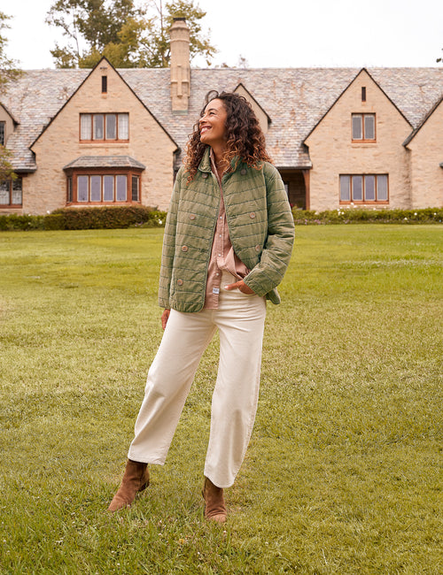 A woman stands on a grassy lawn in front of a large stone house, wearing an olive green quilted jacket over a dusty pink shirt, cream wide-leg pants, and brown ankle boots., view 6