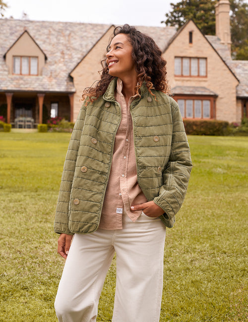 A woman stands on a grassy lawn in front of a large stone house, wearing an olive green quilted jacket over a light pink button-up shirt and off-white pants., front view