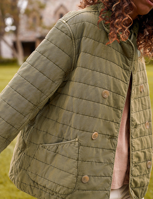 A close-up of a person wearing an olive green quilted jacket outdoors, with grass and a blurred building in the background. The jacket has a button-front closure and a visible pocket., view 5