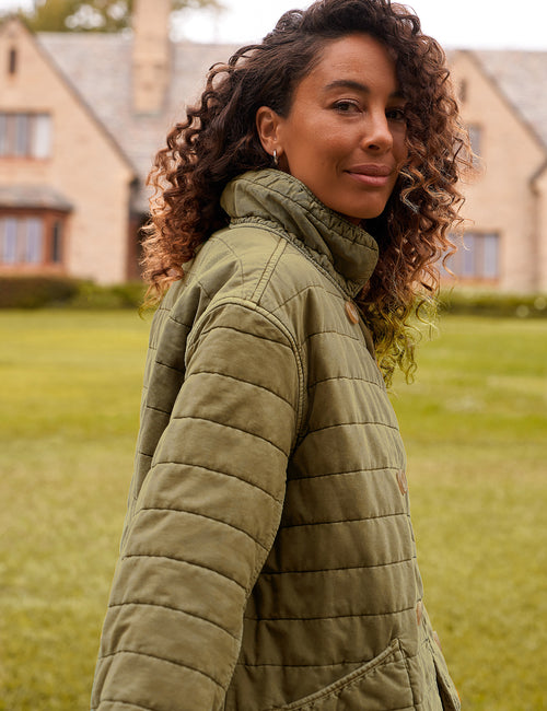 A woman with curly brown hair wearing an olive green quilted jacket, standing outdoors on a grassy lawn with a large stone house in the background., alternate view