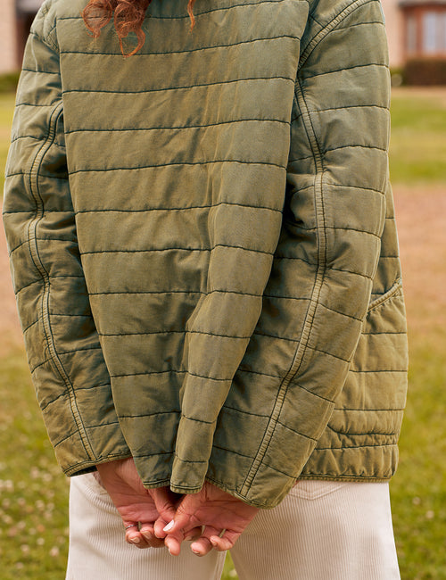 A close-up of a person from behind wearing an olive green quilted jacket and cream-colored pants, standing outdoors on a grassy lawn., view 3