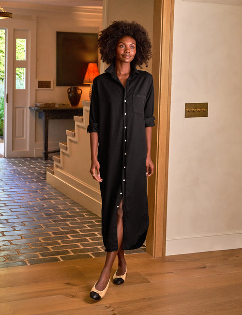 A person standing in an entryway wearing a long black button-up dress with a statement chest pocket, and easy-to-roll sleeves., front view