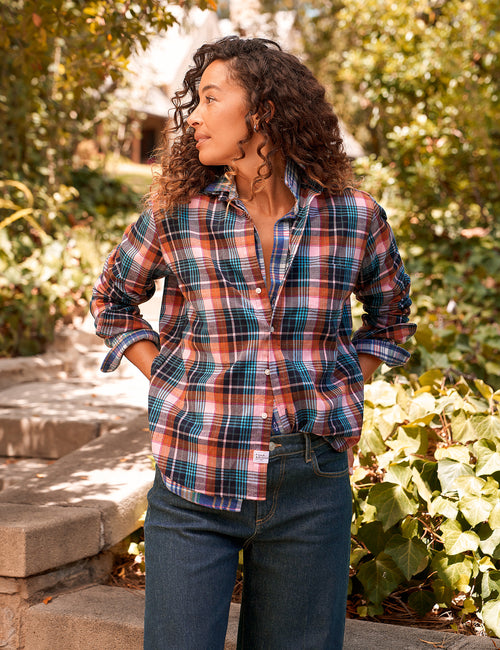 A woman wearing a long-sleeve plaid button-up shirt in pink, blue, and white, paired with dark blue jeans, standing outdoors on stone steps with green foliage in the background., view 4