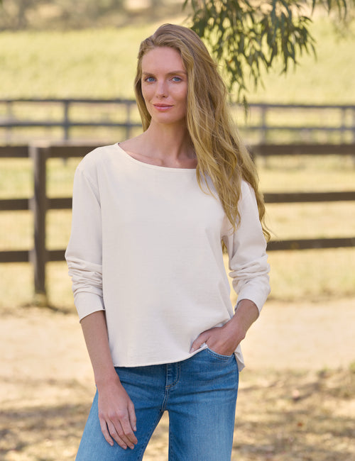 A woman wearing a cream-colored long-sleeve boatneck top and blue jeans, standing in an outdoor rural setting with a wooden fence and greenery in the background., front view