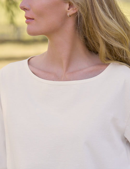Close-up side profile of a woman wearing a cream-colored scoop-neck top and a small gold hoop earring, set against a softly blurred outdoor background., view 3
