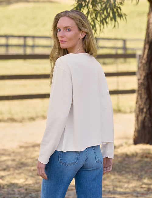 A woman wearing a cream-colored long-sleeve top and blue jeans, photographed from the back in an outdoor rural setting with a wooden fence and tree in the background., alternate view