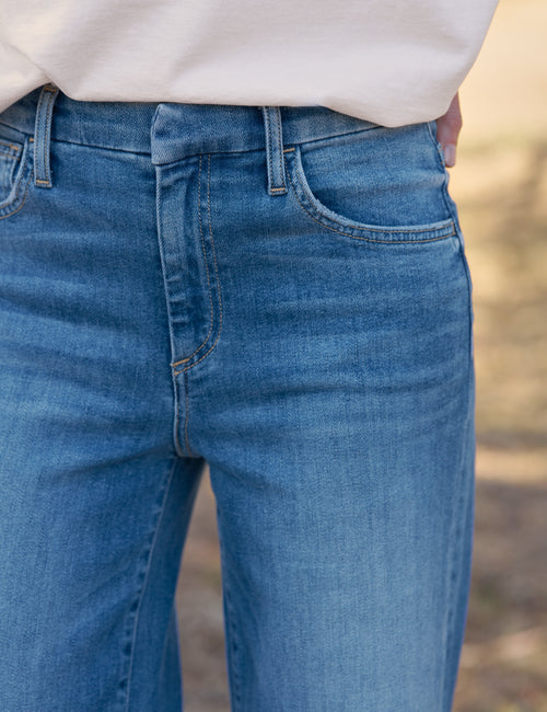 Close-up of a person wearing medium-wash blue denim jeans with a white top visible at the waist, against a softly blurred outdoor background., view 3