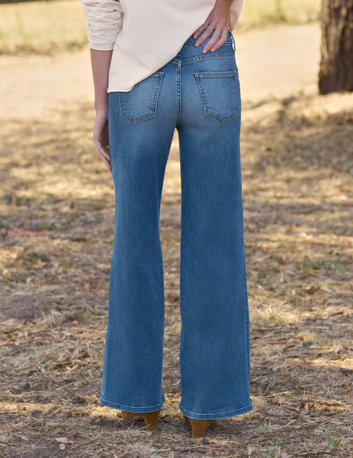 A person shown from the back wearing blue wide-leg jeans and a cream top, standing outdoors on dry grass with trees in the background., alternate view
