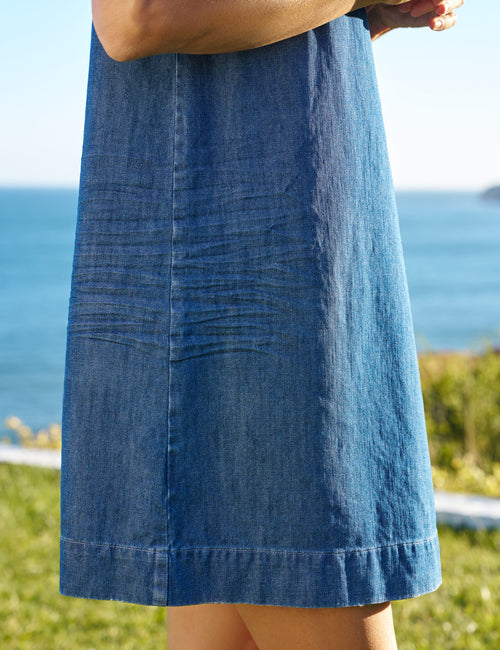 Person wearing a blue denim dress with a blurred background of water and sky, view 4