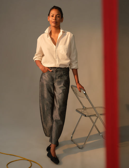 A woman stands in a studio wearing a white button-up shirt and dark gray barrel-leg cropped pants, holding a clear folding chair. A red vertical bar is visible on the right side of the frame., view 4