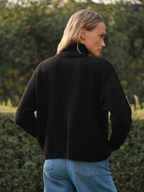 Person wearing a black sweater and blue jeans standing outdoors with greenery in the background, alternate view