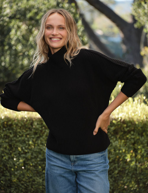 Woman wearing a black sweater and blue jeans standing outdoors with greenery in the background, front view