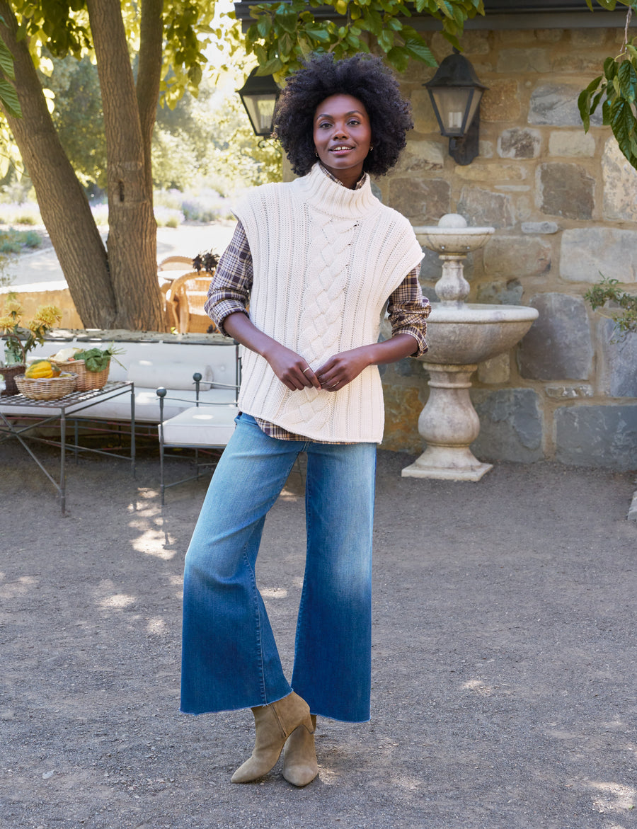 Woman standing outdoors in a garden setting wearing a white cable knit vest, blue jeans, and beige boots.