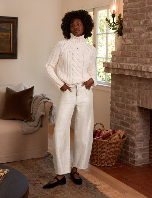 A woman stands in a cozy living room wearing a cream cable-knit mock-neck sweater and matching cream wide-leg trousers, paired with black flats. The room features a brick fireplace, a beige sofa, and a wicker basket., view 4