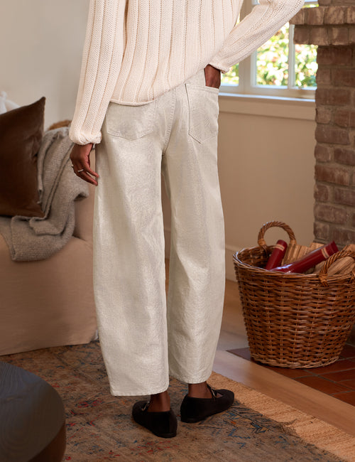 A person wearing loose, barrel-leg cropped pants in a light cream or off-white textured fabric, paired with a ribbed cream sweater and black flats, standing in a cozy indoor room with a couch, basket, and window., alternate view