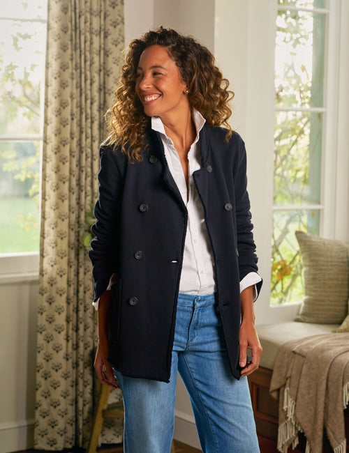 Woman wearing a navy peacoat and blue jeans standing in a bright room with large windows., front view