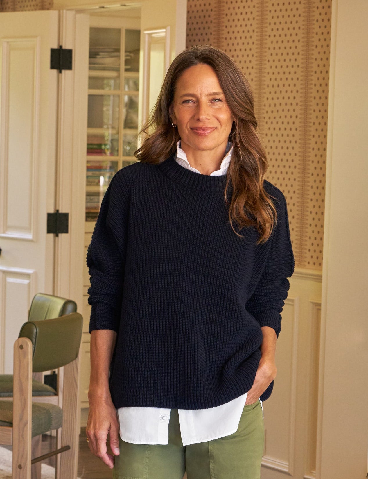 Woman standing in a home interior wearing a navy sweater and green pants.