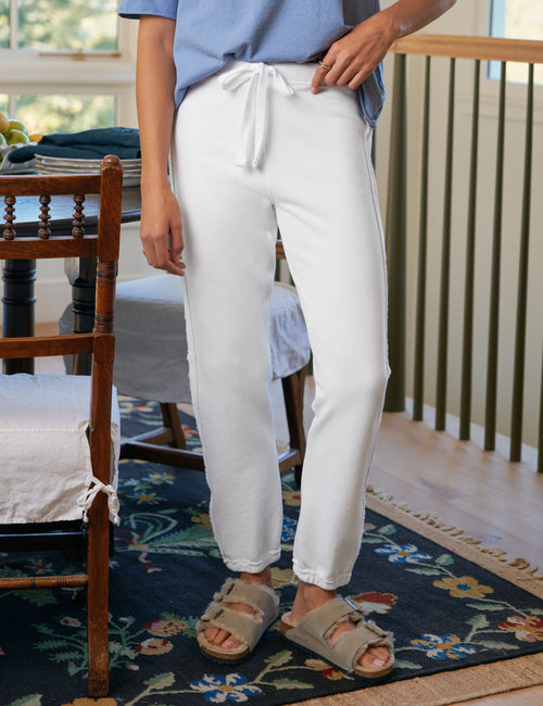 Person wearing white pants and a blue shirt standing in a room with wooden furniture and a floral rug., front view