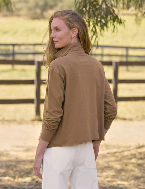 A woman wearing a brown long-sleeve top and white pants, photographed from the back in an outdoor paddock setting with a wooden fence in the background., alternate view