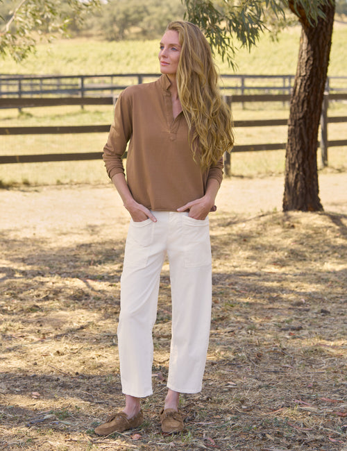 A woman standing outdoors in a sunlit field near a tree and wooden fence, wearing a brown long-sleeve collared henley-style top, off-white cropped pants, and brown slip-on shoes., view 5