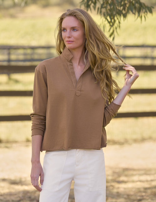 A woman wearing a brown long-sleeve polo-style top and white pants, standing outdoors in front of a blurred wooden fence and dry grass., front view