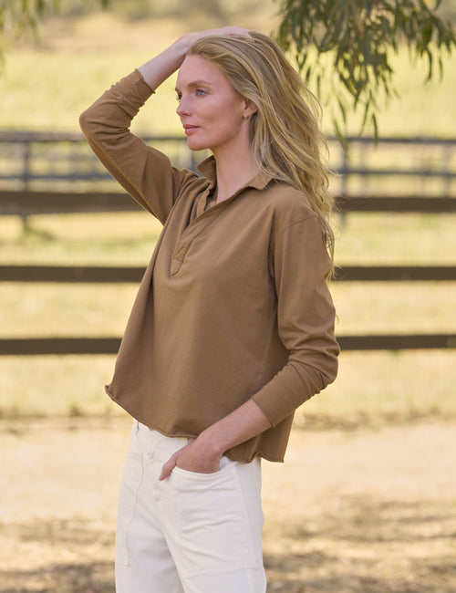A woman wearing a brown long-sleeve collared shirt and white pants standing outdoors in a sunlit field with a wooden fence in the background., view 4