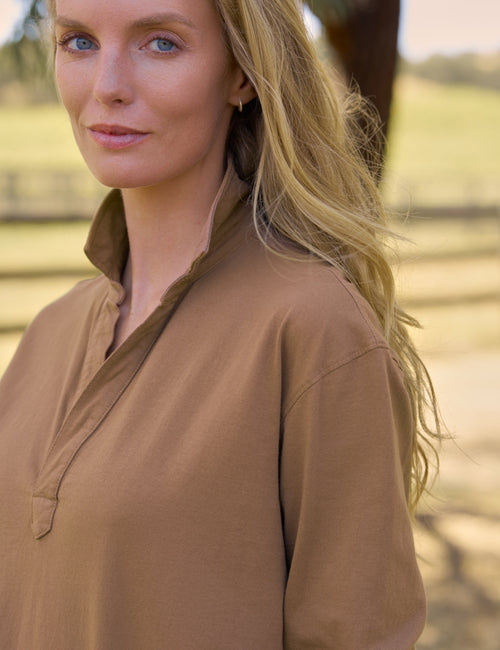 A woman with long blonde hair wearing a brown long-sleeve collared shirt, photographed outdoors against a blurred natural background., view 3