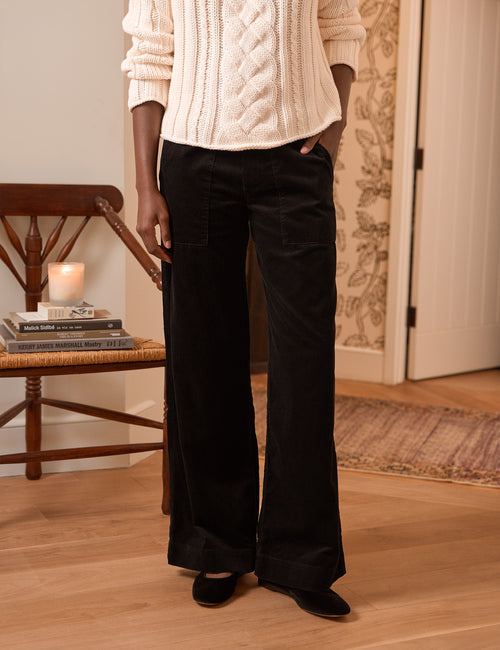 A person wearing black wide-leg corduroy pants with a cream cable-knit sweater, standing in a warm-toned indoor room with a wooden chair and patterned wallpaper., front view