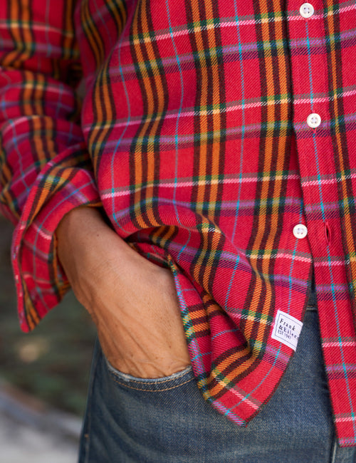 A close-up of a person wearing a pink plaid button-up shirt with blue jeans, showing the fabric texture and a small white brand label near the hem., view 3
