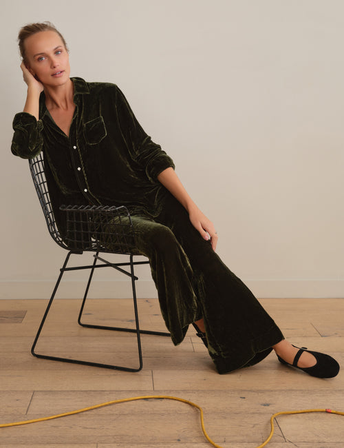 A woman sits on a black wire chair wearing a dark green velvet button-up shirt and matching wide-leg trousers, paired with black flats, in a minimalist room with light wood floors and a beige wall., view 5