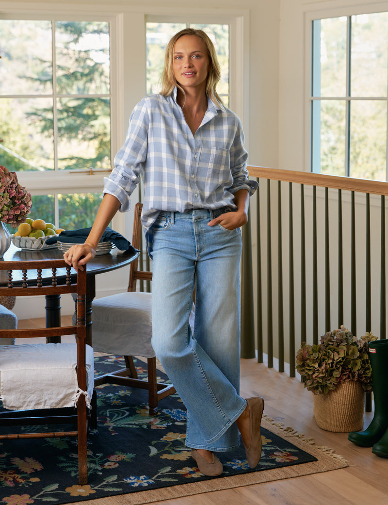 Woman standing in a room with large windows, wearing a checkered shirt and jeans.