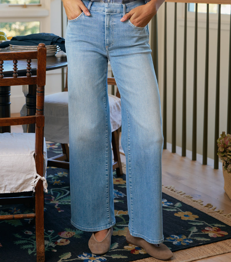 Person wearing light blue jeans standing in a room with wooden furniture and a rug.