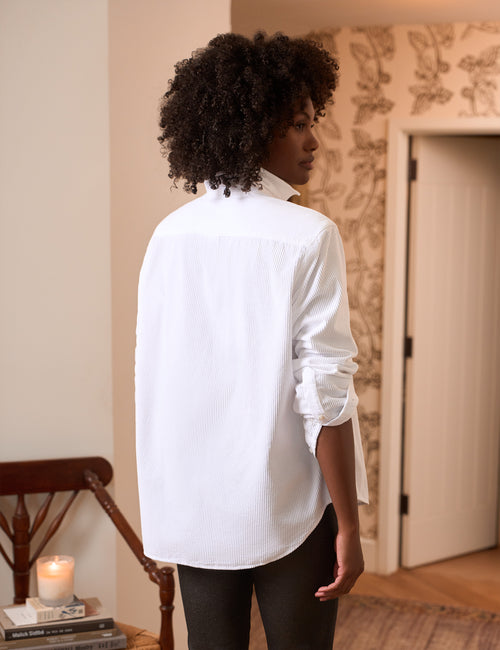 A woman with curly black hair is shown from the back wearing a white button-down shirt and black pants in a warmly lit indoor room with patterned wallpaper and a wooden chair., alternate view