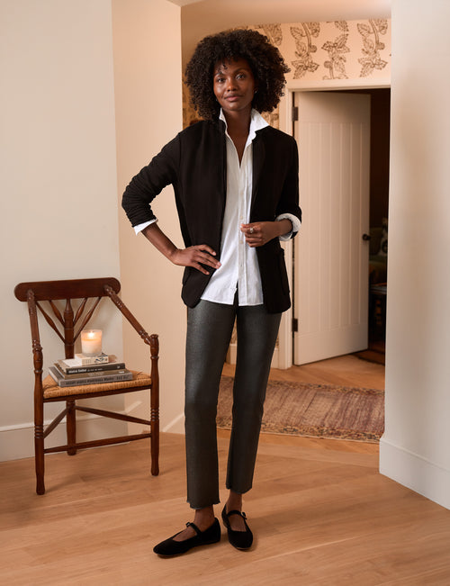 A woman stands in a warmly lit indoor room wearing a black blazer over a white button-up shirt, dark gray cropped pants, and black flats. A wooden chair with books and a candle sits to the left, and an open doorway is behind her., view 4