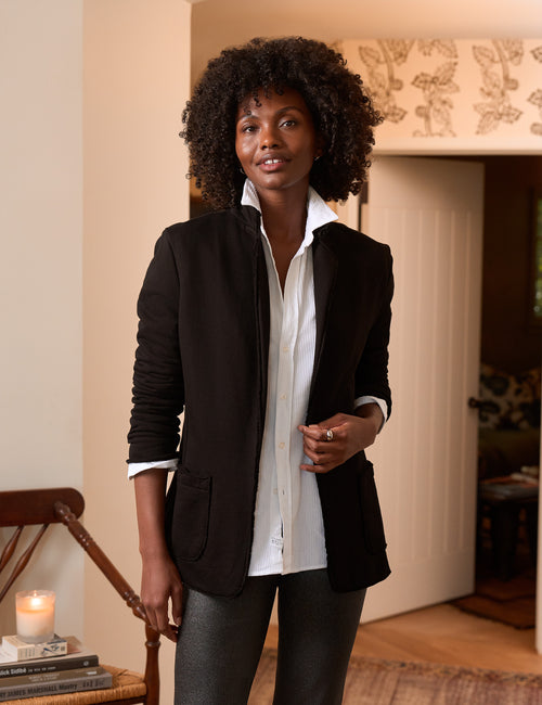 A woman with curly dark hair stands indoors wearing a black blazer over a white button-up shirt and dark gray trousers. The room has warm beige walls, a wooden chair with a lit candle, and a doorway in the background., front view