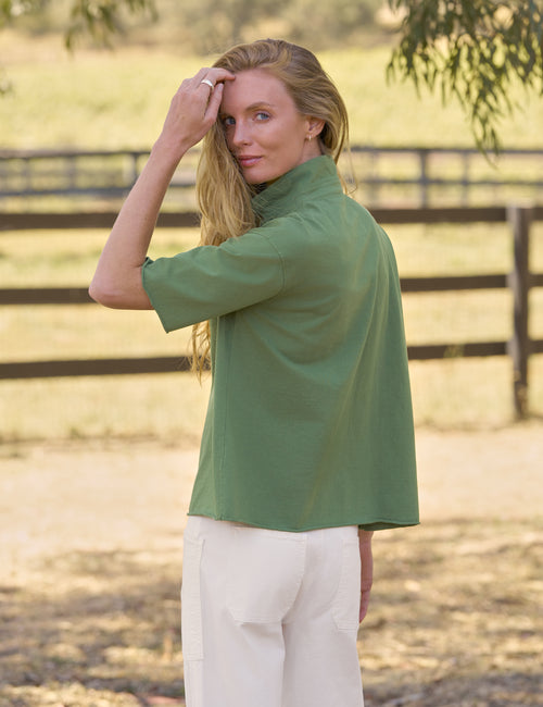 A woman wearing a sage green short-sleeve top and white pants, standing outdoors in a sunlit paddock with a wooden fence in the background., alternate view