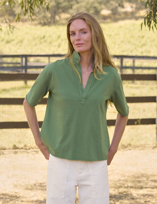 A woman wearing a sage green short-sleeve top and white pants, standing outdoors in front of a wooden fence and grassy field., front view