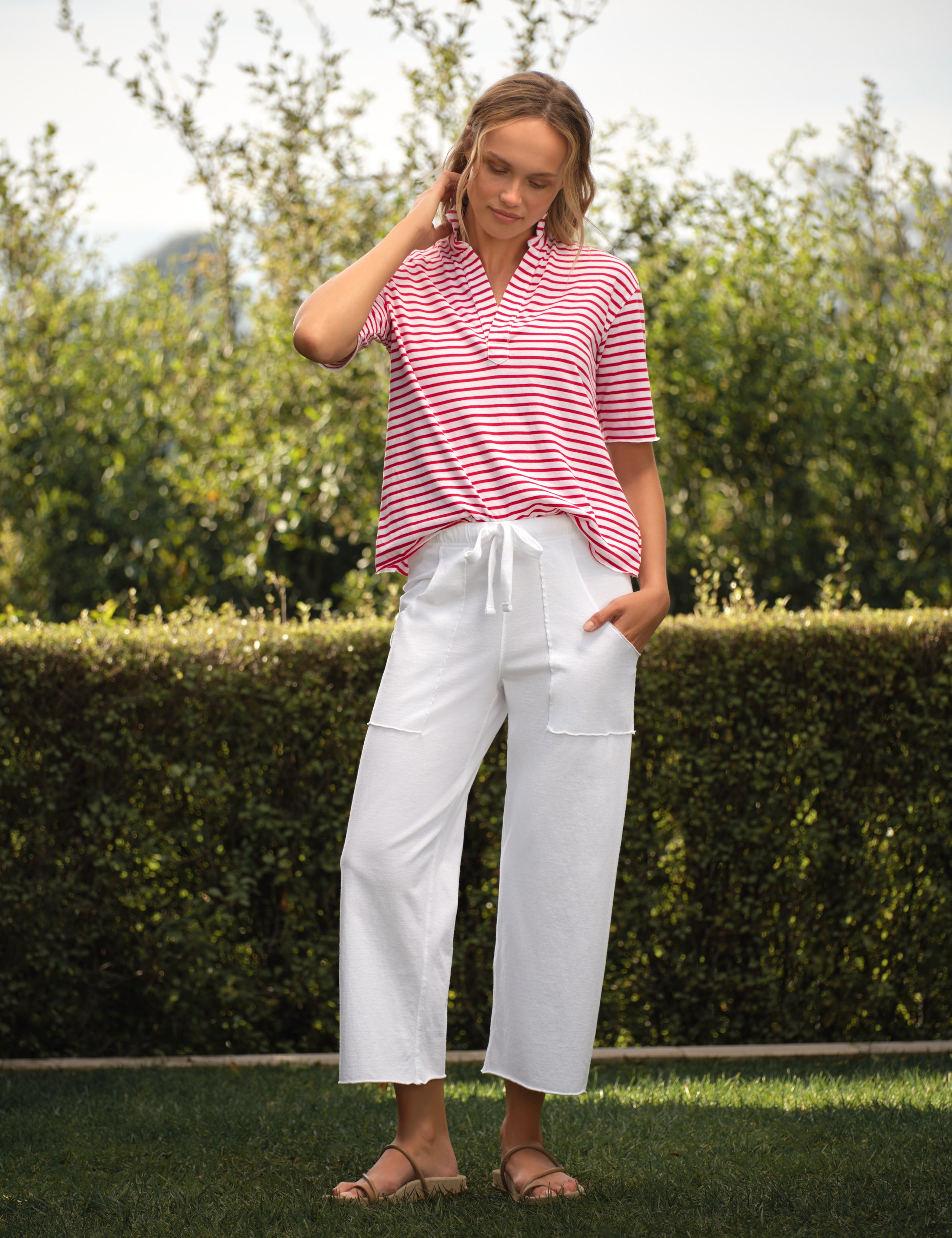 Woman wearing a red and white striped shirt and white pants standing outdoors with greenery in the background, view 4