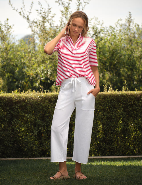 Woman wearing a red and white striped shirt and white pants standing outdoors with greenery in the background, view 4