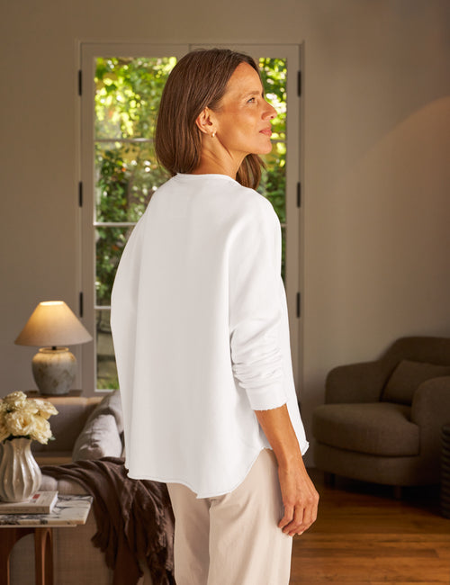 Woman in a white shirt standing in a living room with a view of greenery outside., alternate view
