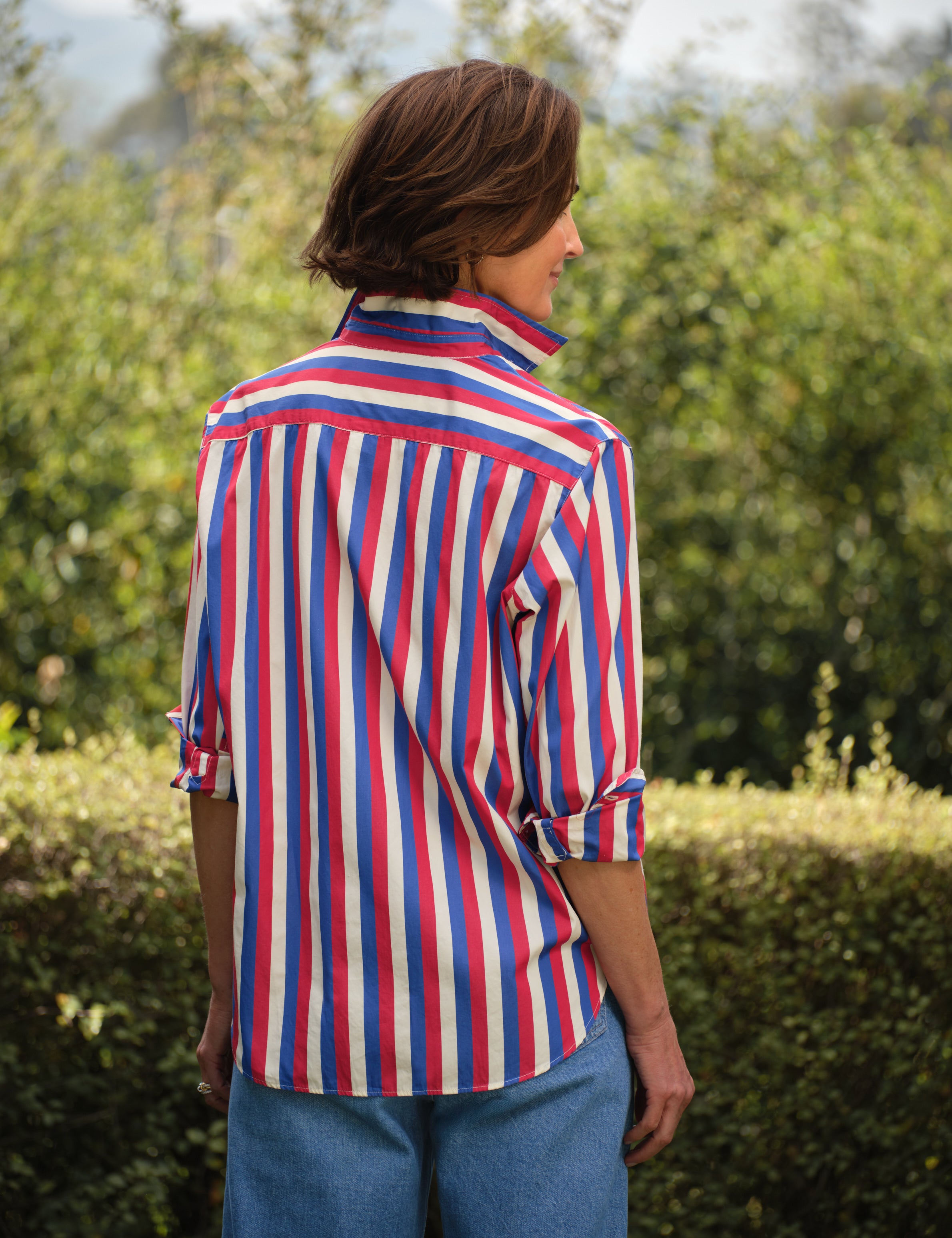 Person wearing a red, white, and blue striped shirt outdoors with greenery in the background, view 4