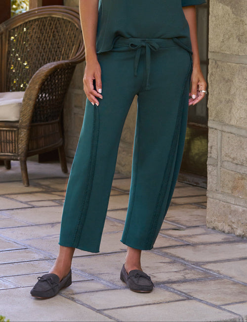 A woman stands on a stone porch wearing a matching dark teal green two-piece lounge set consisting of a short-sleeve crewneck top and wide-leg drawstring pants, paired with dark slip-on shoes., front view