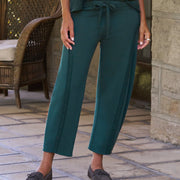 A woman stands on a stone porch wearing a matching dark teal green two-piece lounge set consisting of a short-sleeve crewneck top and wide-leg drawstring pants, paired with dark slip-on shoes.