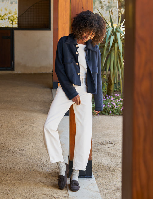 A woman wearing a navy blue cropped jacket, white top, and white wide-leg cropped pants, leaning against a wooden pillar in a modern outdoor corridor., view 3