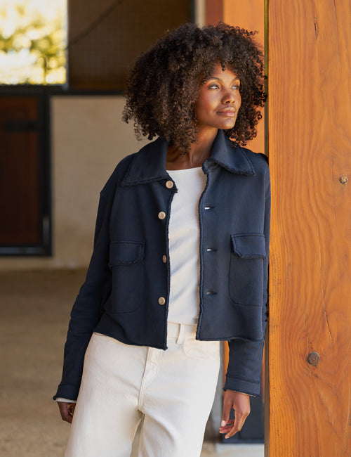 A woman with curly dark hair wearing a navy blue cropped utility jacket over a white top and white pants, standing next to a wooden pillar in an outdoor patio setting., front view