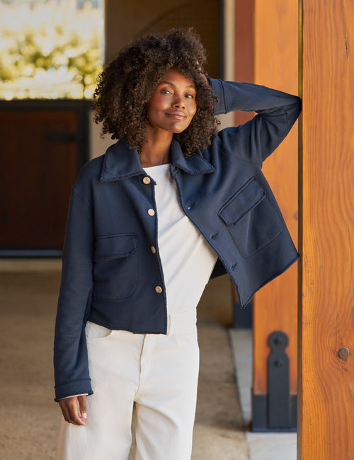 A woman with curly dark hair models a navy blue cropped jacket with gold buttons, worn over a white top and cream pants, leaning against a wooden door., alternate view