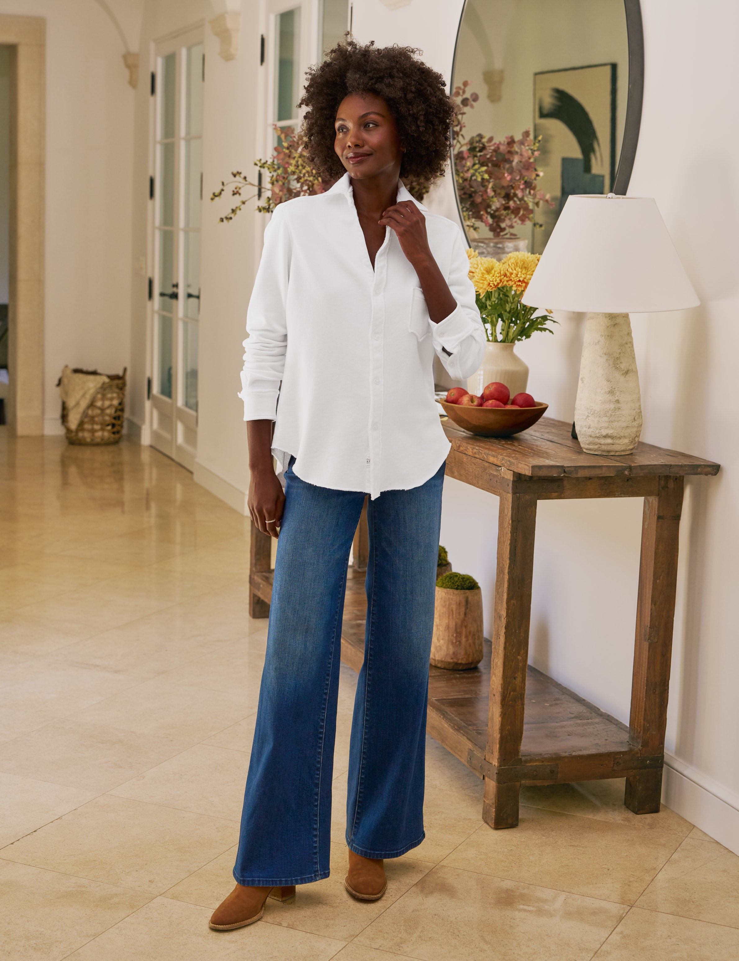 Woman wearing a white shirt and blue jeans standing in a room with a wooden side table and decorative items.