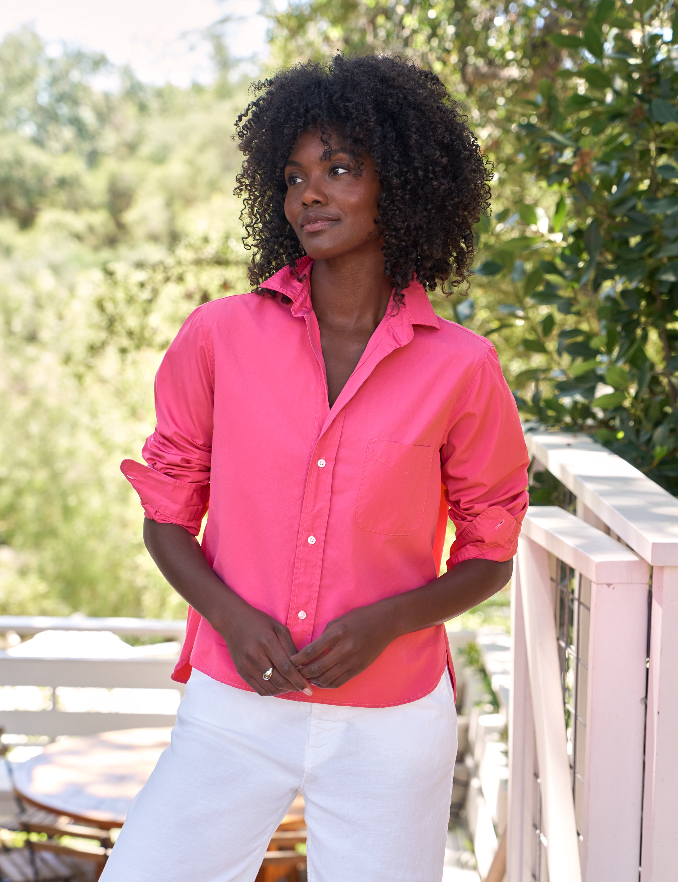 Woman wearing a bright pink shirt and white pants standing outdoors with greenery in the background, view 4